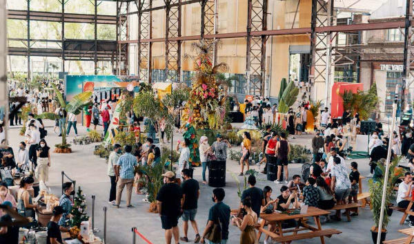 Indoor market scene with many people browsing stalls with plants and food in a large open building with exposed steel beams.